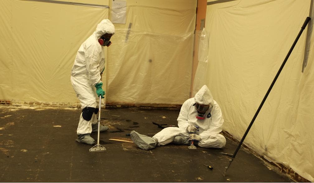 Licensed asbestos abatement crew in Tyvek PPE working under polyethylene containment with negative-air HEPA filtration inside a pre-1980 residence pre-renovation removal scene (Wikimedia Commons, CC BY 2.0)