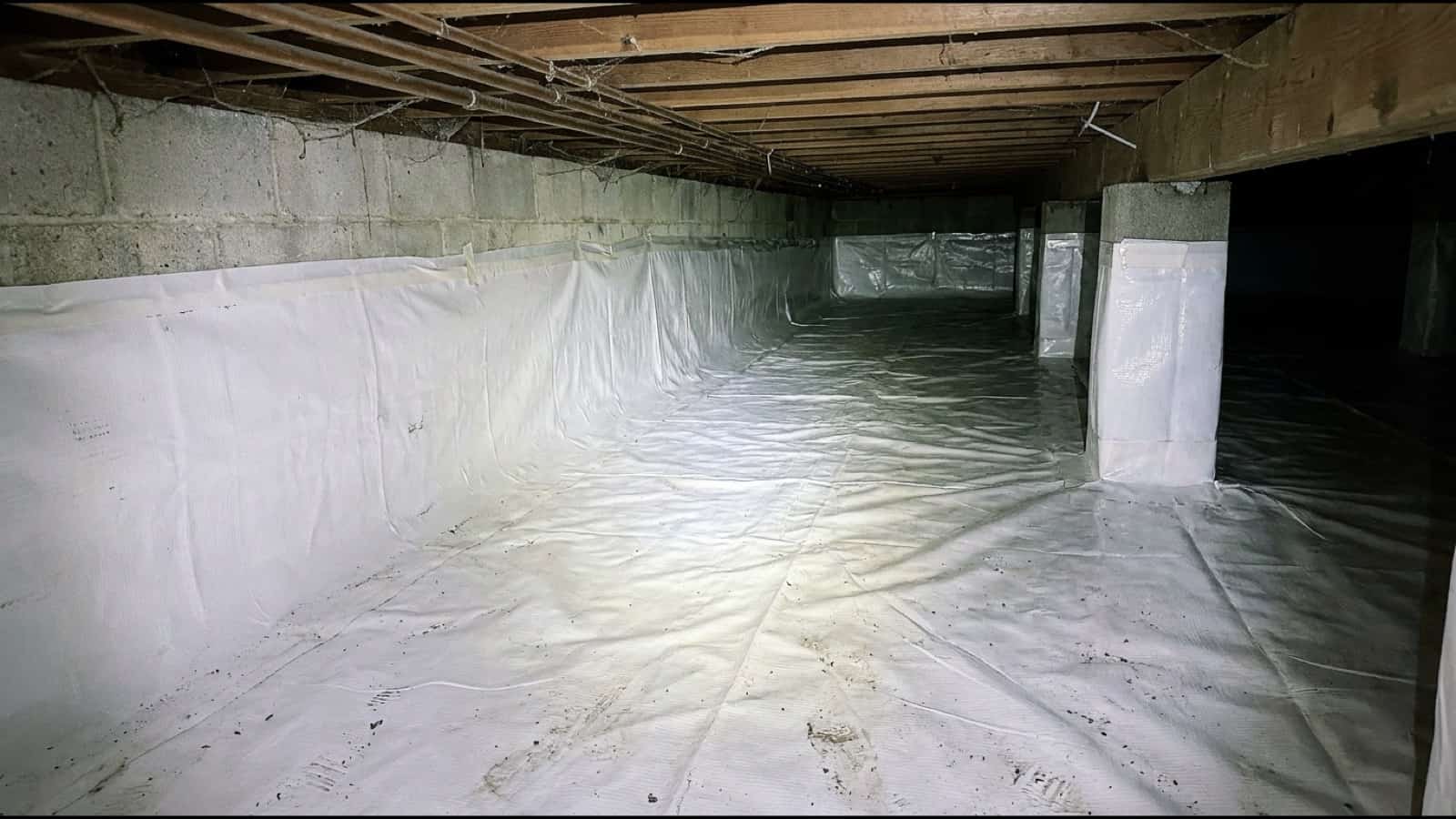 Finished crawl space encapsulation in a Connecticut home: white sealed liner across the floor and up the block wall, wrapped piers, photographed by phone flash