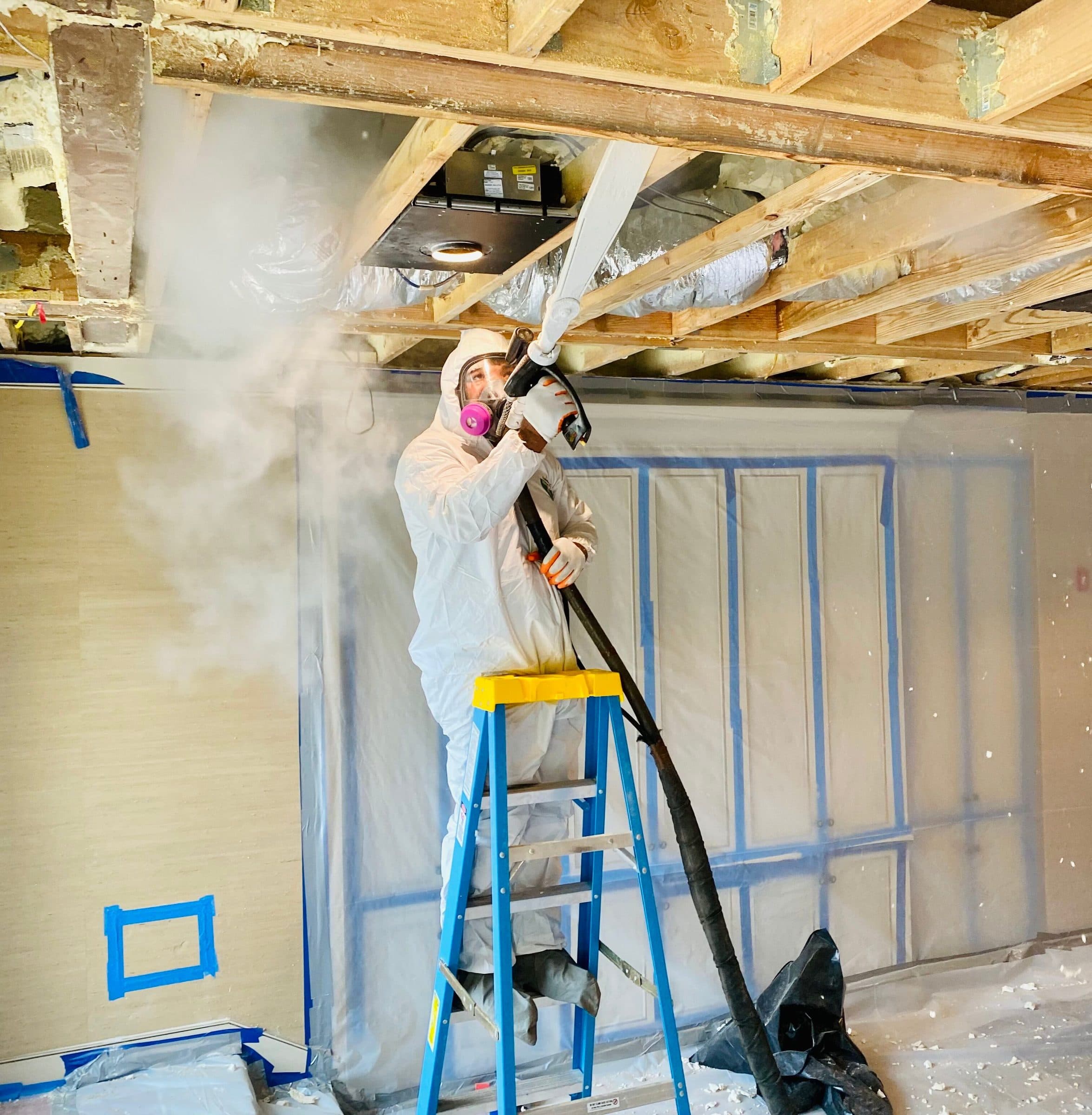 Green Restoration technician in full Tyvek PPE and respirator treating an attic with visible black mold across the roof sheathing and rafters during active IICRC S520 mold remediation