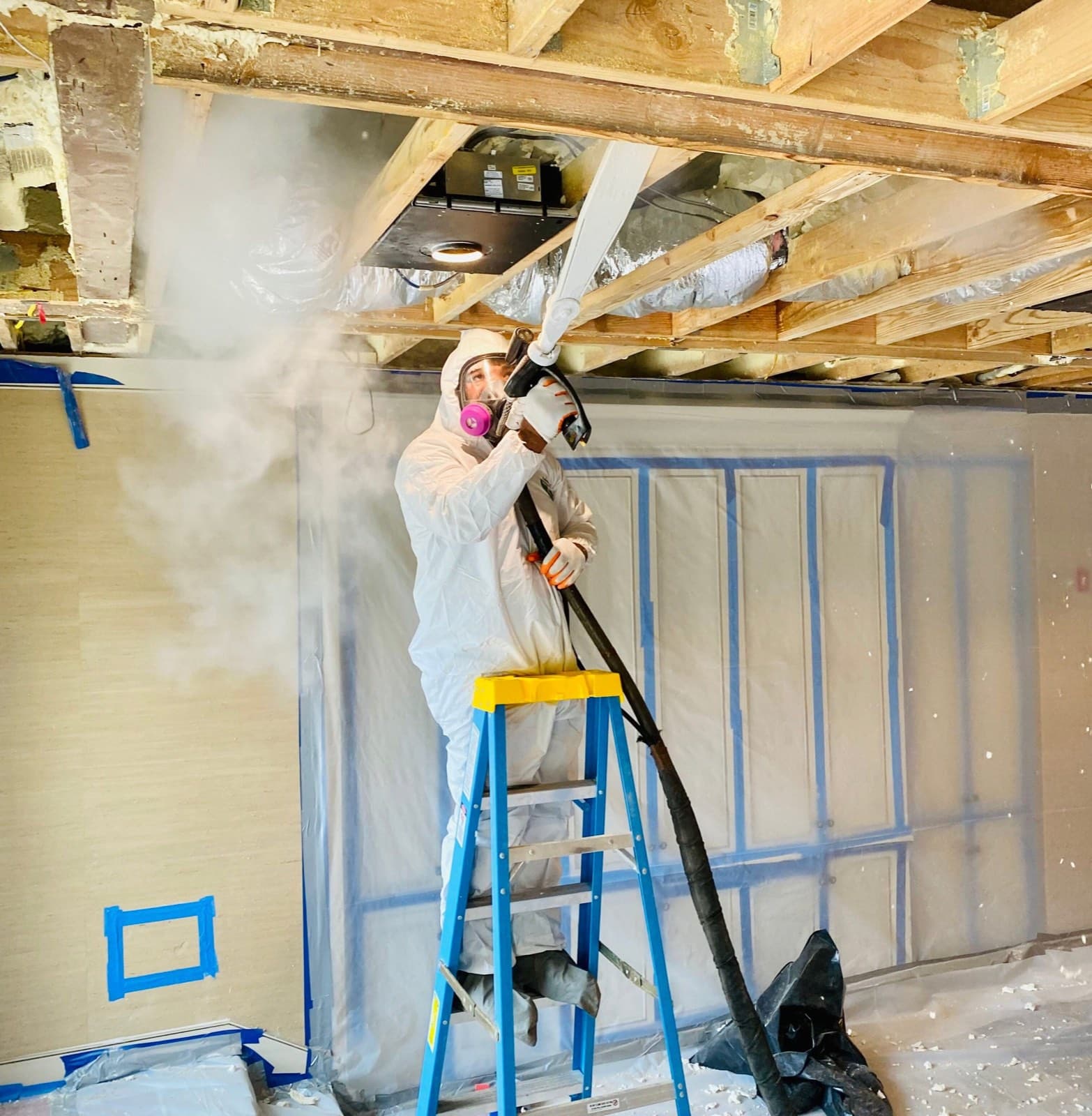 Green Restoration technician in full Tyvek PPE and respirator on a ladder treating a residential attic with visible mold growth across the roof sheathing and rafters during an active IICRC S520 remediation