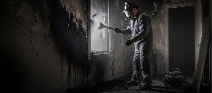 IICRC S700 technician wearing a respirator and nitrile gloves wiping heavy black soot residue from a painted interior wall with a dry chemical sponge, test swabs and a HEPA vacuum staged on a drop cloth below