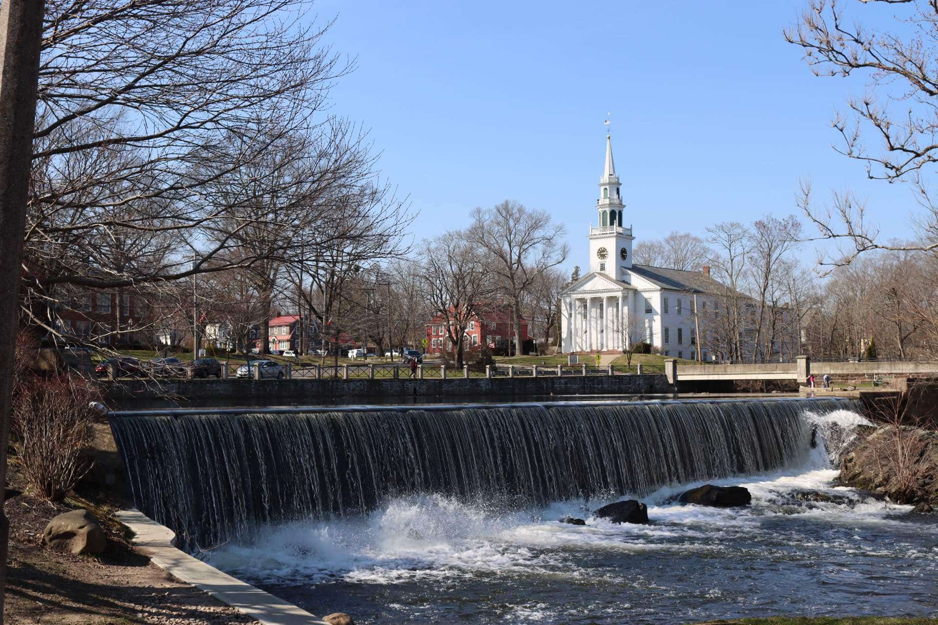 Milford, Connecticut duck pond waterfall with the First Congregational Church steeple. Green Restoration provides 24/7 water, fire, mold, and storm damage restoration in Milford, CT.