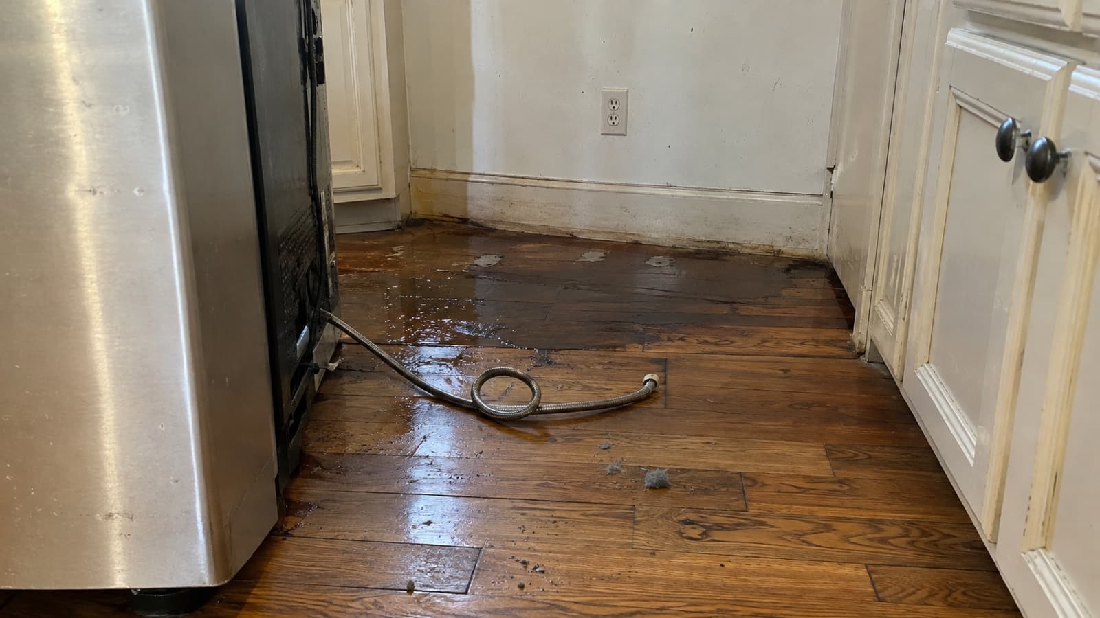 Refrigerator rolled out of its kitchen alcove with a braided ice maker water line lying in a puddle on darkened hardwood floor boards