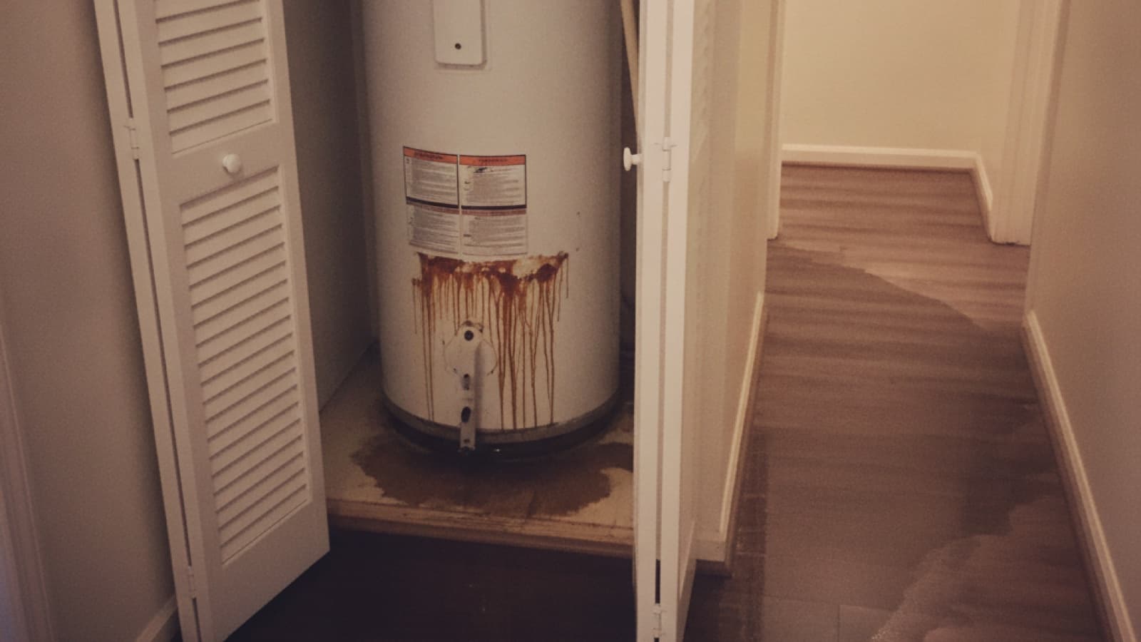 Rust-streaked water heater in an open hallway closet with leaked water spreading over the threshold across the wood floor